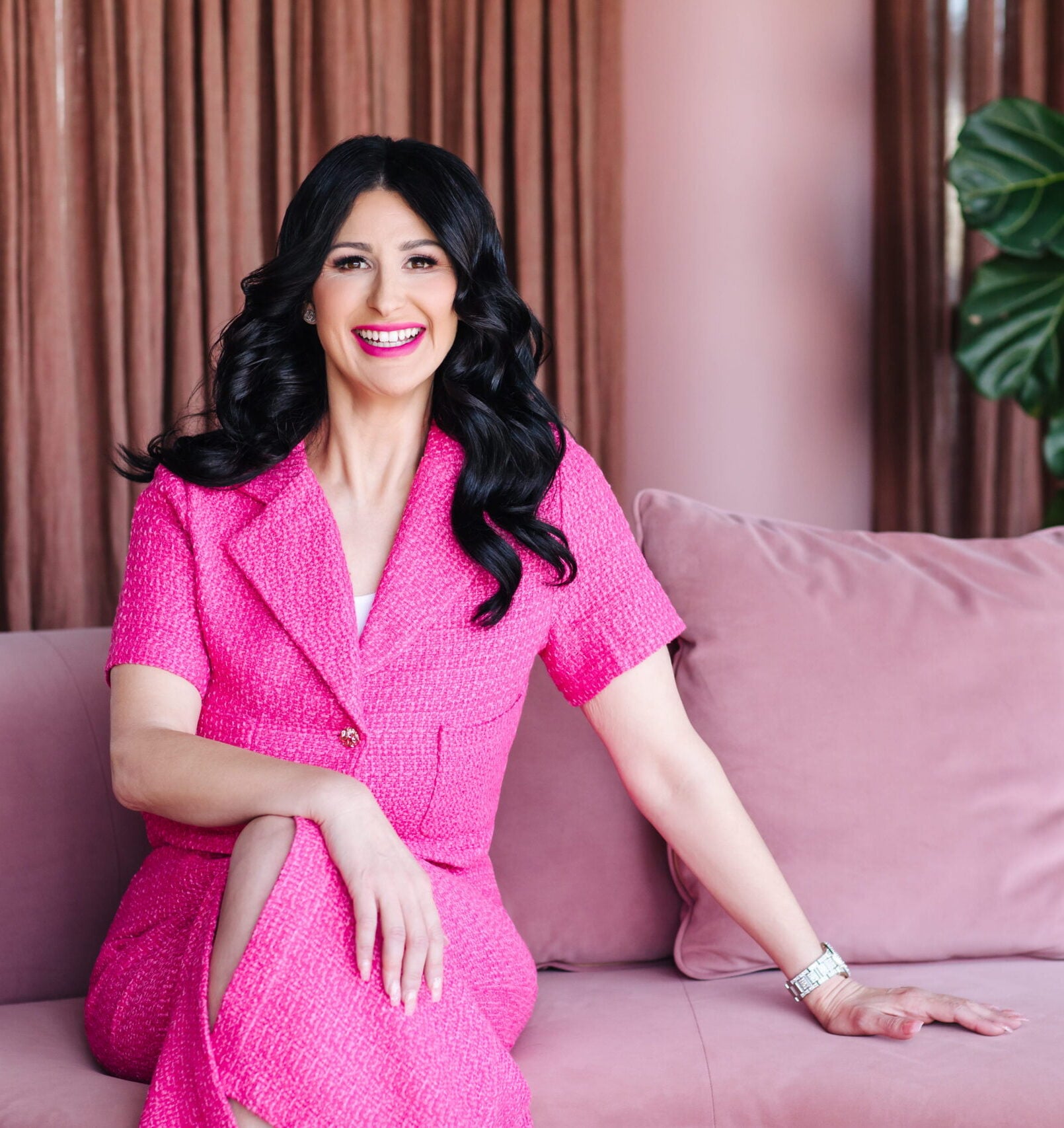 Gee, Melbourne wedding planner and founder of Gee | What a Wedding | smiling confidently in a pink tweed blazer and skirt set, seated on a blush pink sofa in a stylish pink-toned studio setting