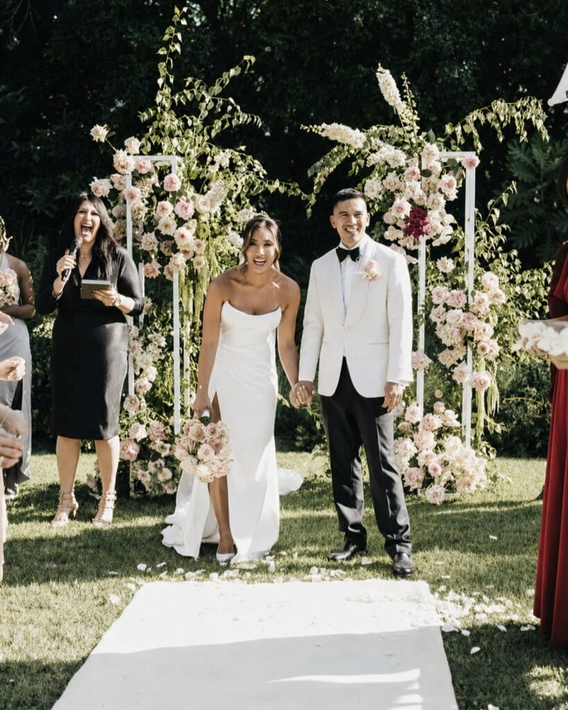 Bride and groom walking a confetti-covered aisle with Melbourne marriage celebrant Gee Tankir cheering, ceremony at a private park in Melbourne's southeastern suburbs, floral installations either side